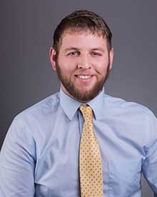 A headshot of a friendly-looking man with a short beard and brown hair, sporting a light blue shirt with a yellow tie adorned with blue polka dots. He has a cheerful expression and stands against a gray backdrop.