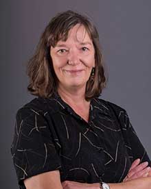 A headshot of a woman with a warm smile, medium-length brown hair, and earrings. She is wearing a dark blouse with a unique twig and splinter pattern in lighter tones, her arms crossed comfortably. The background is a smooth, neutral gray.