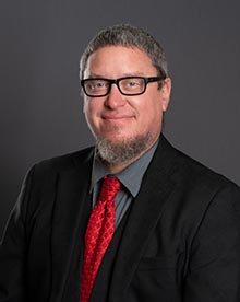 A professional headshot of a smiling man with graying hair and a beard, wearing black-framed glasses, a dark gray suit, a light gray shirt, and a red tie with a diagonal square pattern.