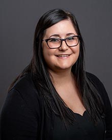 a portrait of a woman with long black hair with black rimmed glasses. she has a happy smile and is wearing a professional black top