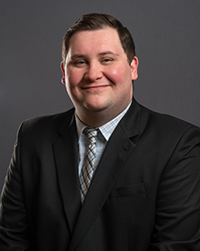 A professional headshot of a man with a content smile, short dark hair, wearing a black suit, a white shirt with a light blue checkered pattern, and a diagonally striped brown and white tie. The backdrop is a dark gray.