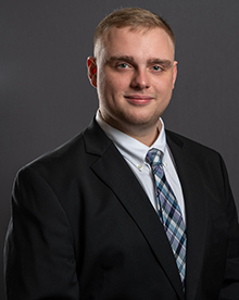 a man with short blond hair wearing a black suit with a white shirt and blue plaid patterned tie with a confident smile