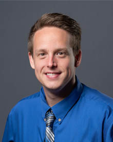 a professional portrait of a man with short brown hair with a big smile. He is wearing a blue shirt with a blue and black striped tie.