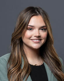 a professional headshot of a woman with long brown hair wearing a green jacket and black shirt with a warm smile. The background is dark gray.