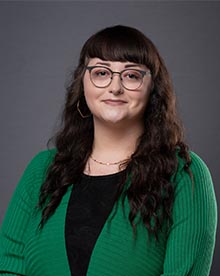 a woman with long dark hair wearing dark glasses. she has a content smile and is wearing a knit sweater and black shirt