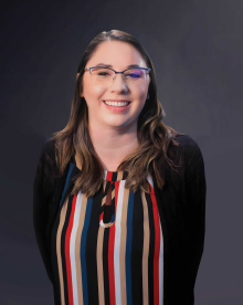 a woman with a big smile and long wavy hair and thin rimmed glasses wearing a professional striped shirt and cardigan sweater