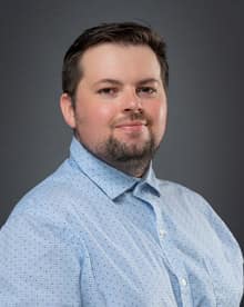 professional photo of a man with brown hair and brown facial hair wearing a blue button up shirt