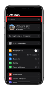 Namedrop the image shows an iphone with its settings screen open. At the top is the search bar, used for quickly finding settings. Below the search bar, there's a section with an icon representing the "1st National Bank", suggesting that the bank's app is integrated with the device's settings, possibly for Apple ID, iCloud, media, and purchases. Other visible settings options include "Get Help During an Emergency", "AirPods Pro" listed as "1NB", indicating a custom naming for the device, and standard settings such as "Airplane Mode", "Wi-Fi", "Bluetooth", "Cellular", "Personal Hotspot", "Notifications", and "Sounds & Haptics". The focus of the image seems to be on the integration and ease of access to the 1st National Bank settings within the device's system settings