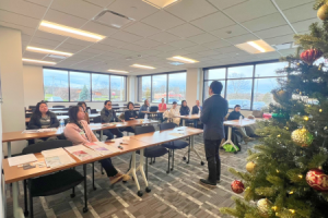 The image captures a professional setting where a business seminar or workshop is taking place. A speaker is standing at the front of a room filled with attentive participants seated at tables. The audience, consisting of a diverse group of adults, appears to be engaged in the presentation. Natural light streams in from the windows, creating a bright and inviting atmosphere. To the right, a festively decorated christmas tree adds a seasonal touch to the room, suggesting the event is taking place around the holiday season. The setting conveys a sense of community and learning, with a focus on interaction and education.