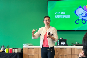 The image shows a woman presenting in front of a screen with a vibrant green background. She appears to be in the midst of explaining a concept, with her hand raised in a gesture that indicates she's making a point. The screen behind her displays "2023" prominently, along with Chinese characters, suggesting the topic relates to plans or strategies for the year 2023. The presentation seems to be taking place in a casual setting, with a coffee station and supplies visible in the background, indicating a relaxed yet professional atmosphere.