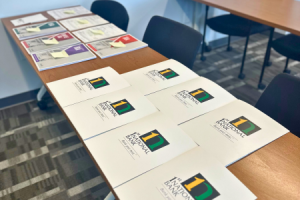 The image shows a long table in a conference room setting, laid out with informational packets. The packets are neatly arranged in rows, with the top visible page featuring a colorful logo and the text "1st national bank," indicating the material is likely related to the bank's services or the event being held. The room is well-lit and has a modern, professional ambiance with a clean and organized look, suggesting a preparation for a seminar or workshop where participants will receive these materials.