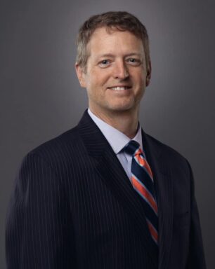 Professional headshot of a man in a navy pinstripe suit and striped tie, smiling against a dark gray backdrop. Mark Swartz commercial lender 1st National Bank